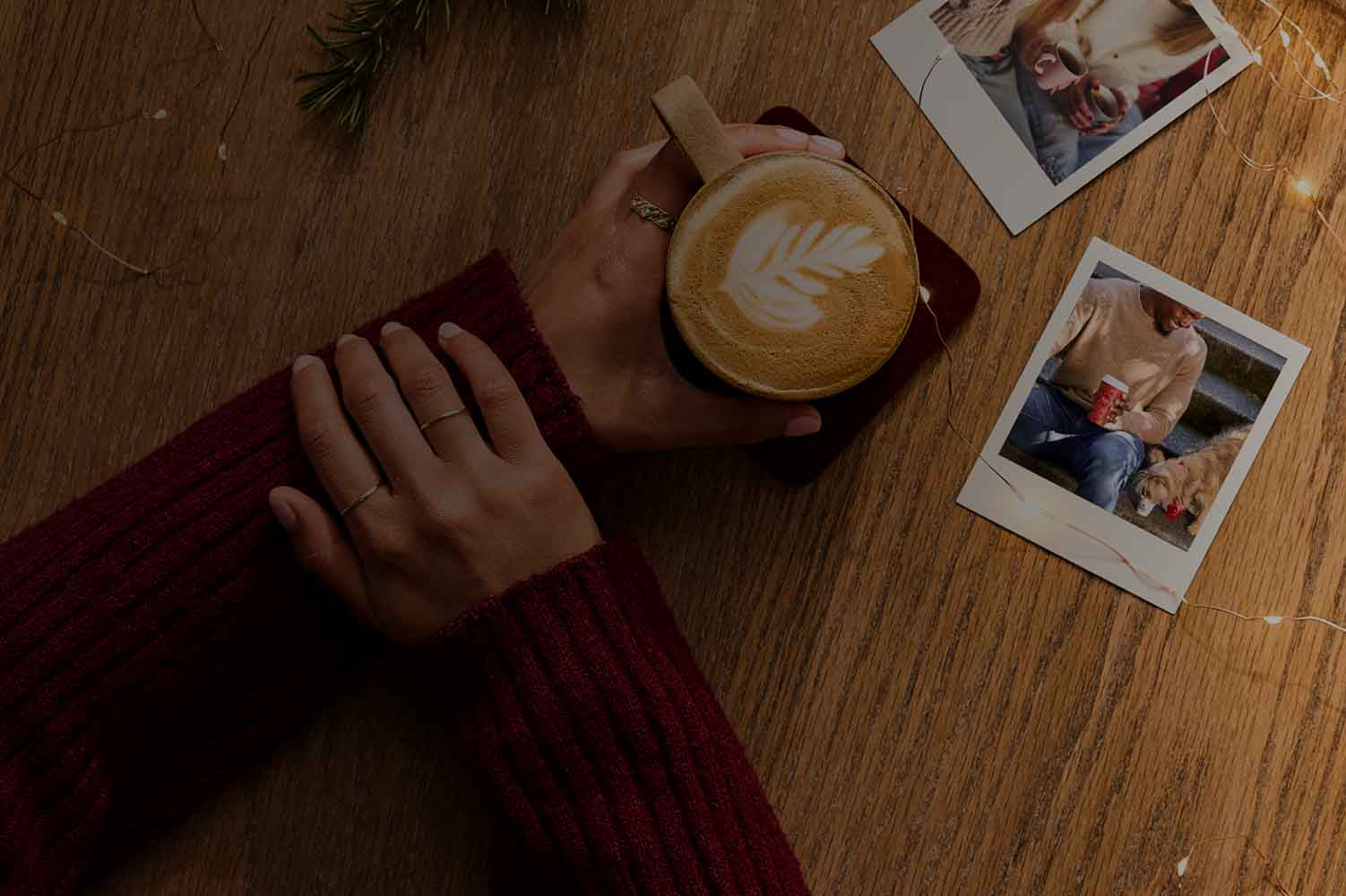 Person holding a coffee cup with latte art on a wooden table with photos and a red blanket.