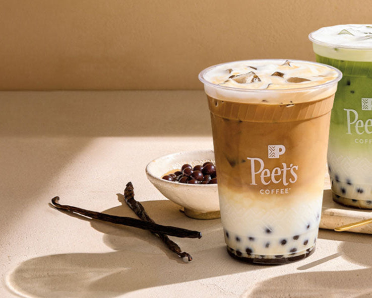 Two to-go Peet's cups both with coffee pearls at the bottom on a counter with a vanilla bean.