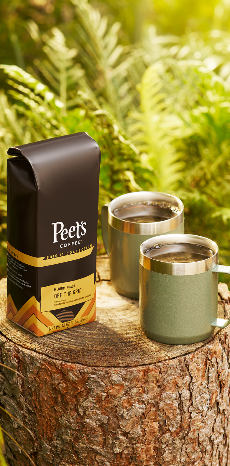 Bag of coffee beans next to two green travel mugs on top of a tree stump in the forrest