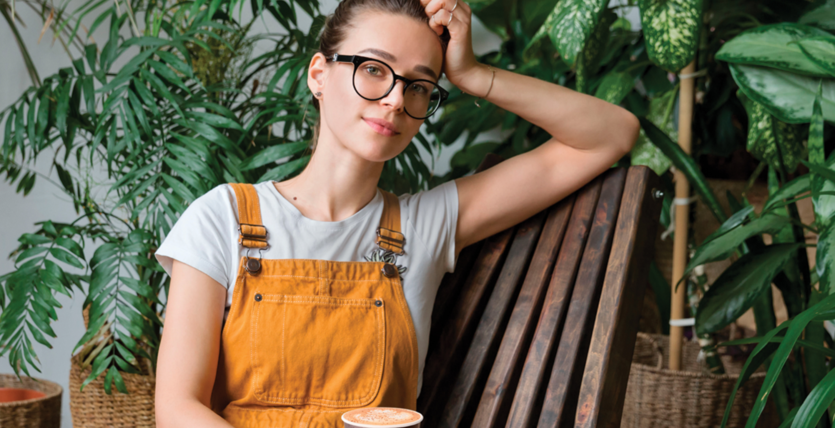Women in orange overalls sitting in front of plants holding cup of coffee