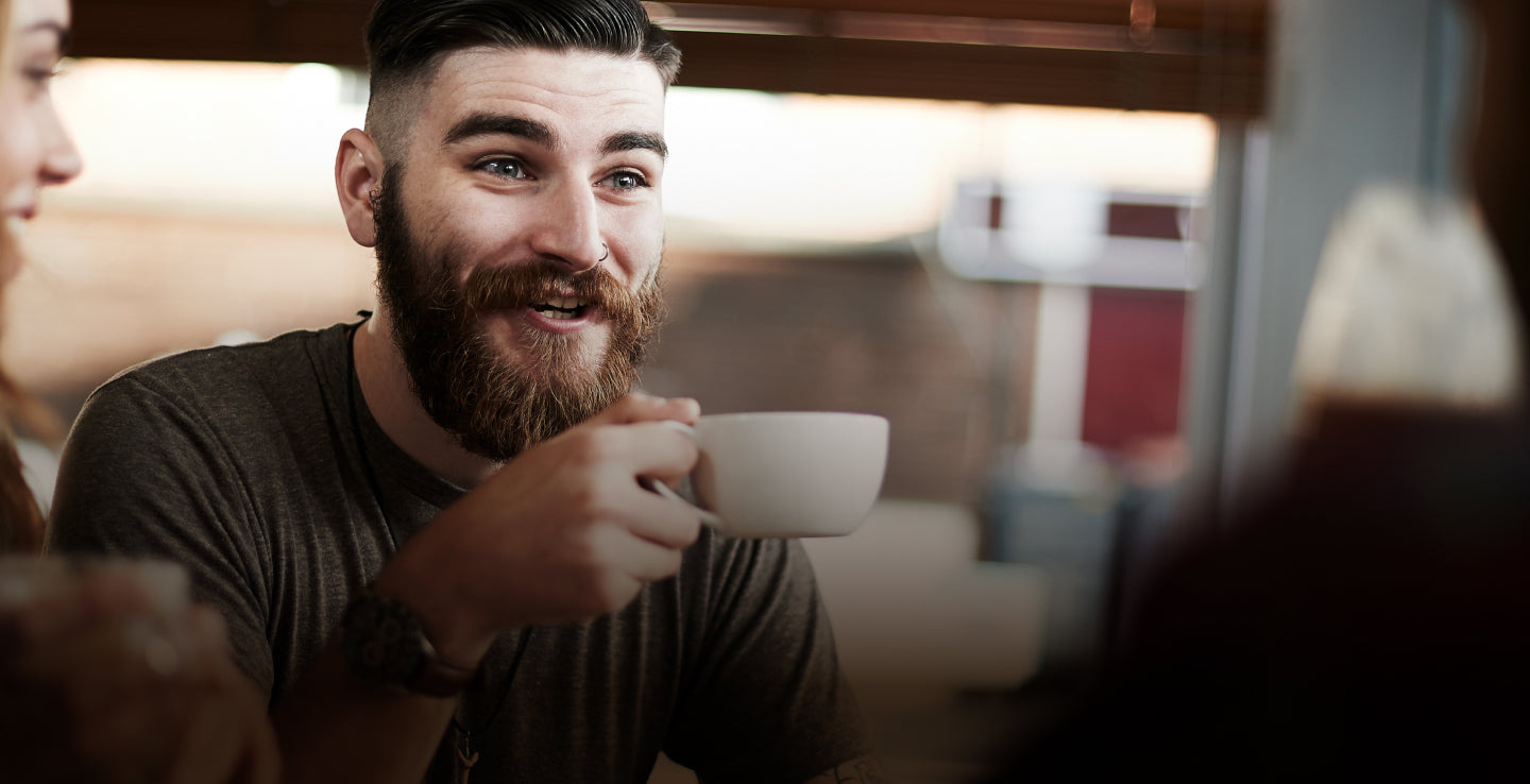 Man with beard holding white mug of coffee talking to friends