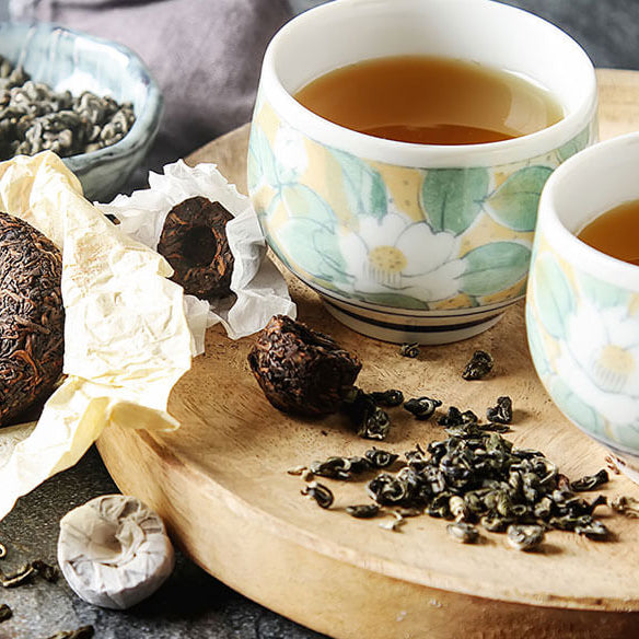 Two cups of tea on a wooden board with tea leaves and a tea bag.